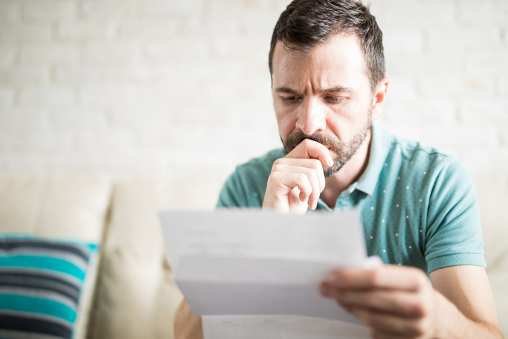 Homem de barba sentado no sofá, olhando para um papel.