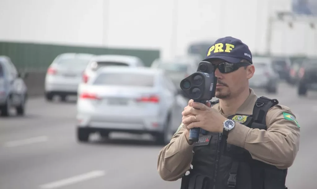 Agente da Policia Rodoviário Federal com um radar móvel na mão apontando para carros na estrada.