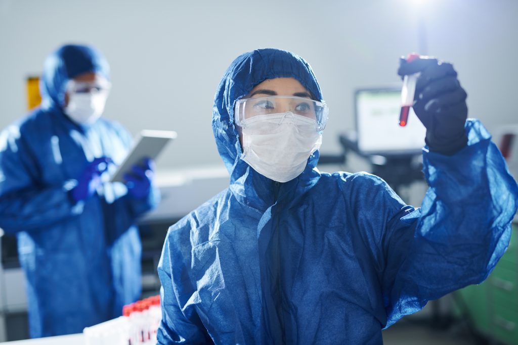 Cientista de saúde em traje de proteção azul de biohazard e óculos de segurança, de pé no laboratório e trabalhando com uma amostra de sangue.