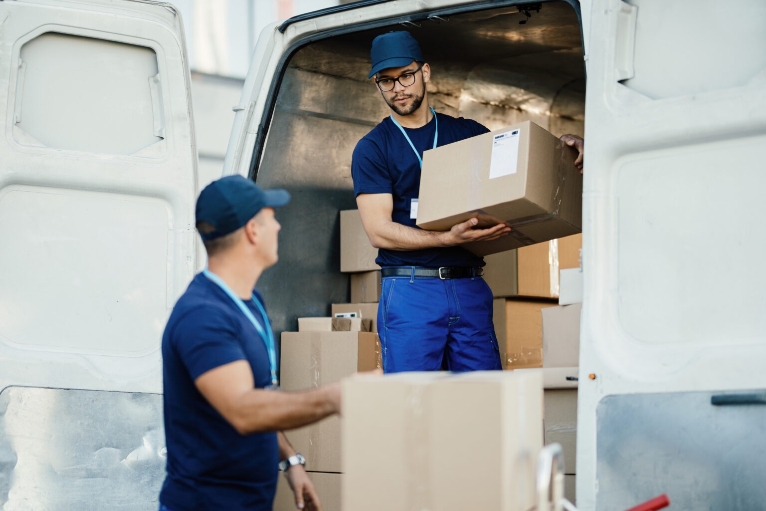 Dois entregadores de uniforme azul carregando caixas de papelão na traseira aberta de uma van branca, um passando a caixa para o outro