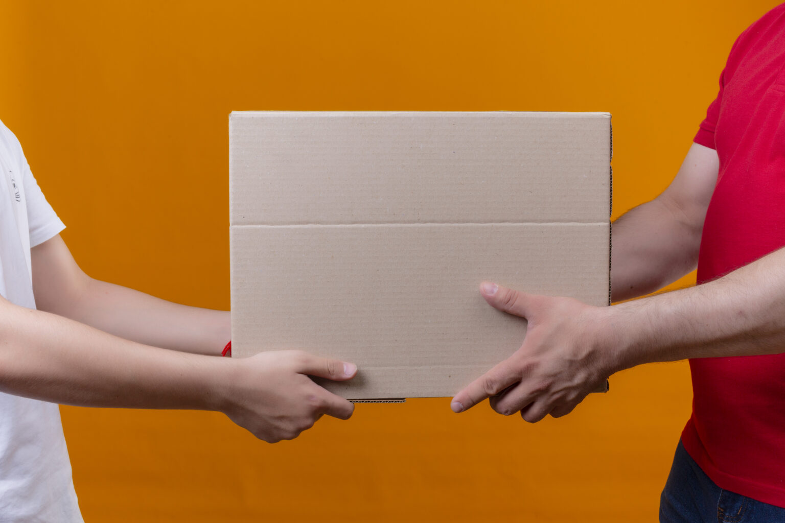 Duas pessoas segurando juntas uma caixa de papelão grande e retangular, vista de frente, contra um fundo laranja liso