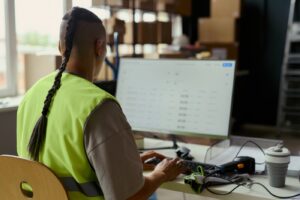 Operador de armazém, visto de costas, consultando uma tabela na tela do computador, com leitor de código de barras sobre a mesa