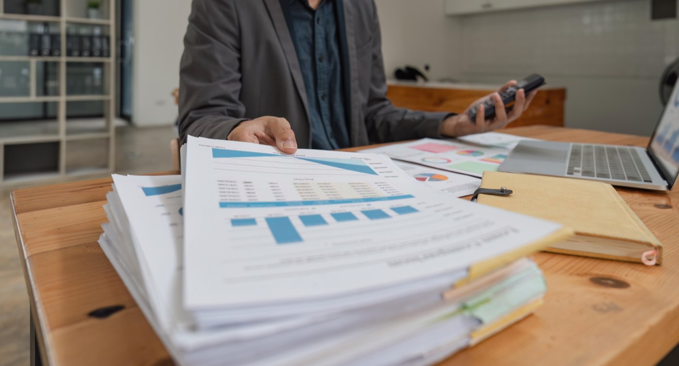 Pilha de relatórios impressos com gráficos sobre a mesa, ao lado de calculadora e notebook