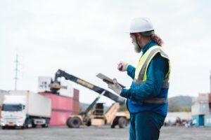 Trabalhador de capacete e colete refletivo consulta um tablet no pátio, com caminhão de baú e contêineres empilhados ao fundo