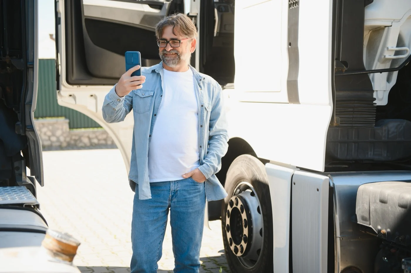 Motorista de barba grisalha, de óculos e camisa jeans, em pé entre duas cabines de caminhão estacionadas no pátio, olhando a tela do celular