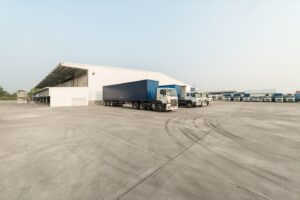 Pátio de transportadora com uma carreta sider engatada e uma fileira de caminhões estacionados em frente ao galpão
