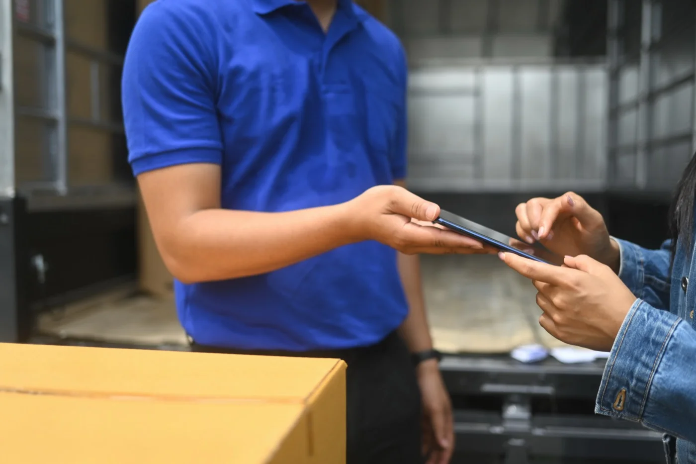Entregador de camisa azul, diante do baú aberto do caminhão, estende o celular para a recebedora assinar na tela, com uma caixa de papelão em primeiro plano