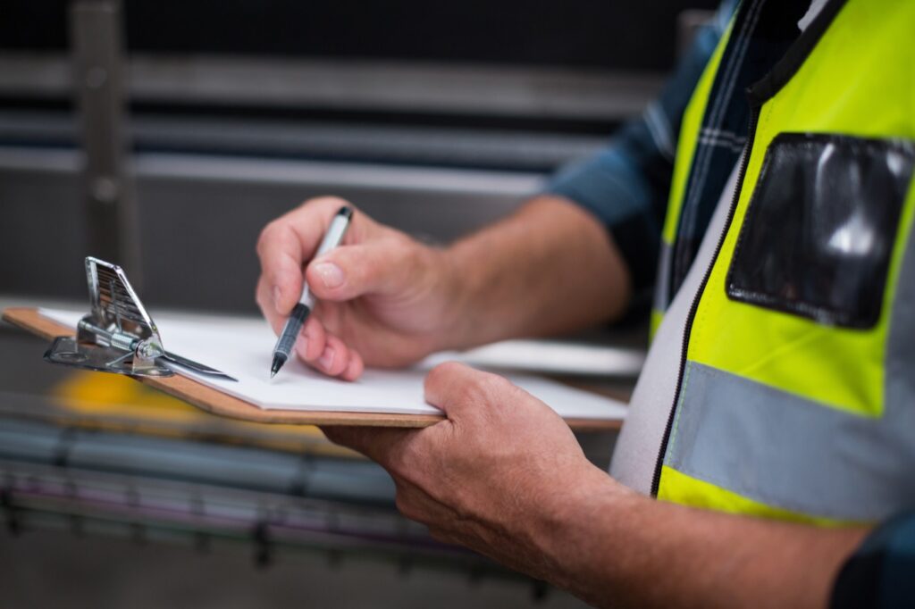 Mãos de um operador de colete refletivo anotando em uma prancheta, com o documento preso pelo grampo