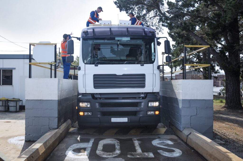 Caminhão parado sobre a balança de um posto de fiscalização, com STOP pintado no chão e dois fiscais conferindo a carga em cima do veículo