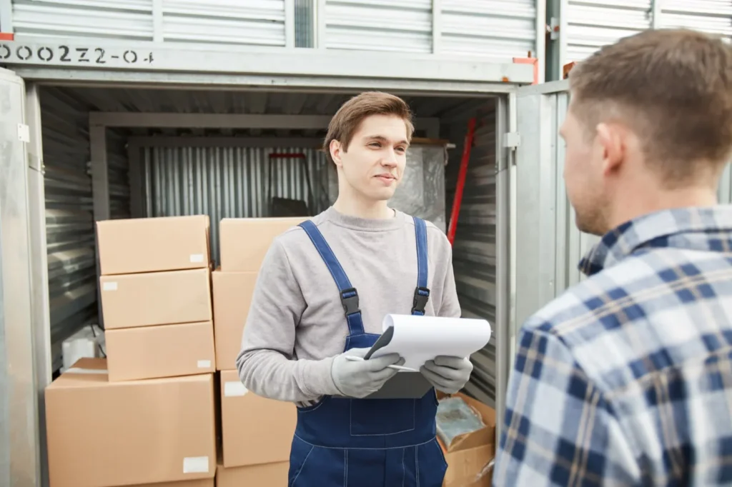 Funcionário de macacão azul e luvas segura uma prancheta e conversa com um cliente de camisa xadrez diante de um contêiner aberto com caixas de papelão