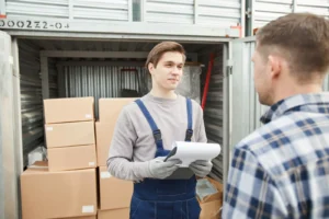 Funcionário de macacão azul e luvas segura uma prancheta e conversa com um cliente de camisa xadrez diante de um contêiner aberto com caixas de papelão
