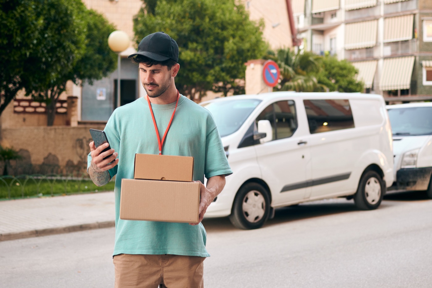 Entregador de camiseta verde segurando duas caixas e consultando o celular na rua, com vans brancas estacionadas ao fundo