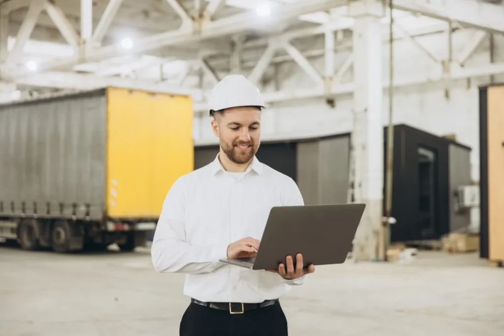 Homem de capacete branco e camisa social sorri enquanto usa um notebook, com uma carreta de lona amarela e cinza ao fundo, em um galpão amplo