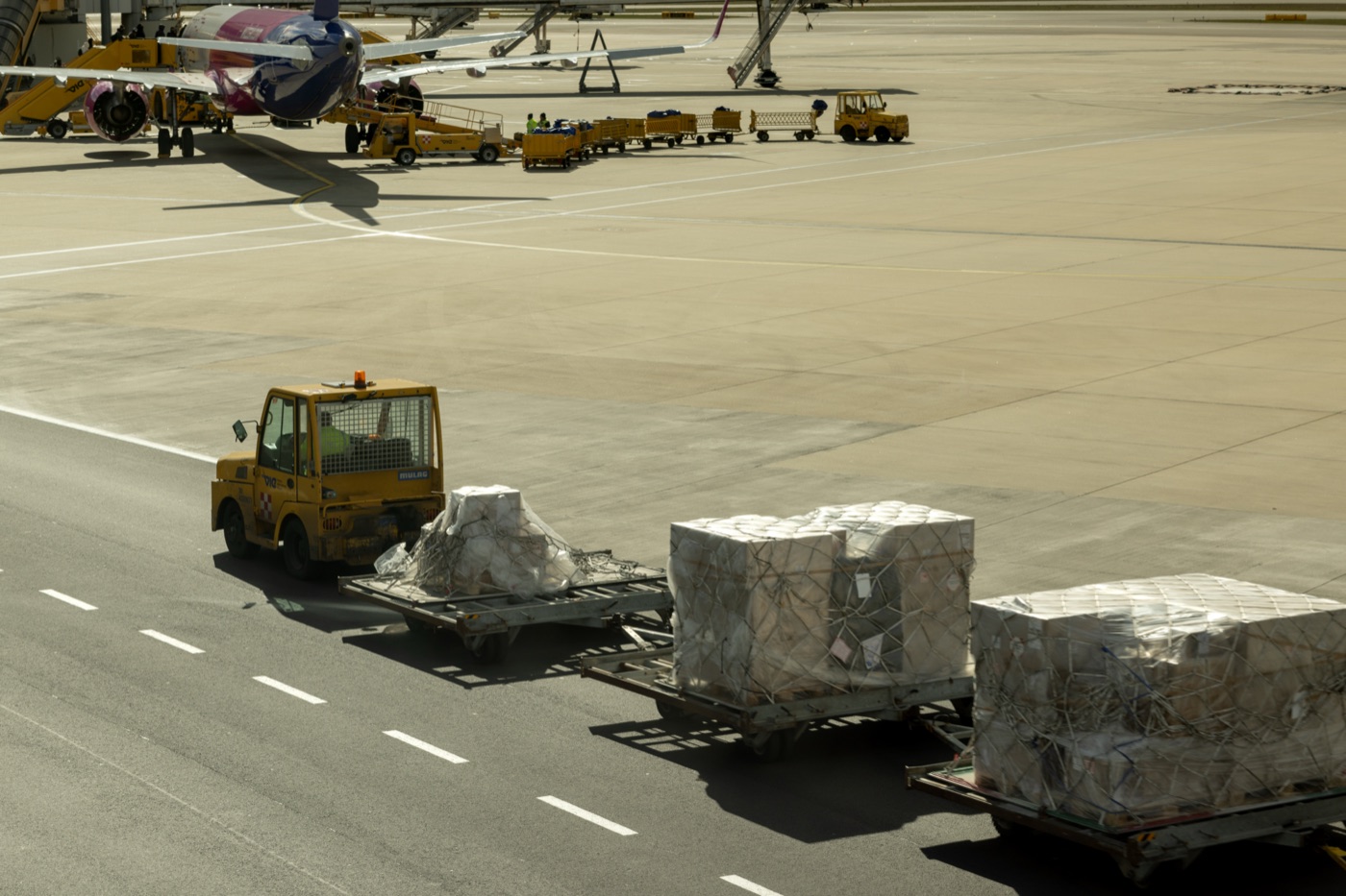 Trator de pátio puxando três paletes de carga aérea embalados em rede na pista do aeroporto, com aviões ao fundo
