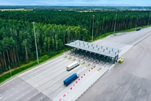 Vista aérea de uma praça de pedágio em rodovia cercada por floresta, com dois caminhões se aproximando das cabines